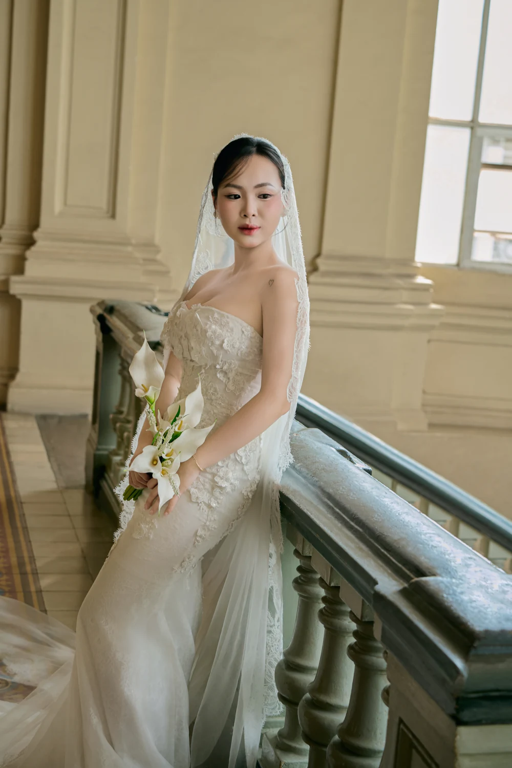 The bride on the staircase, calla lilies gathered against her lace, glancing away